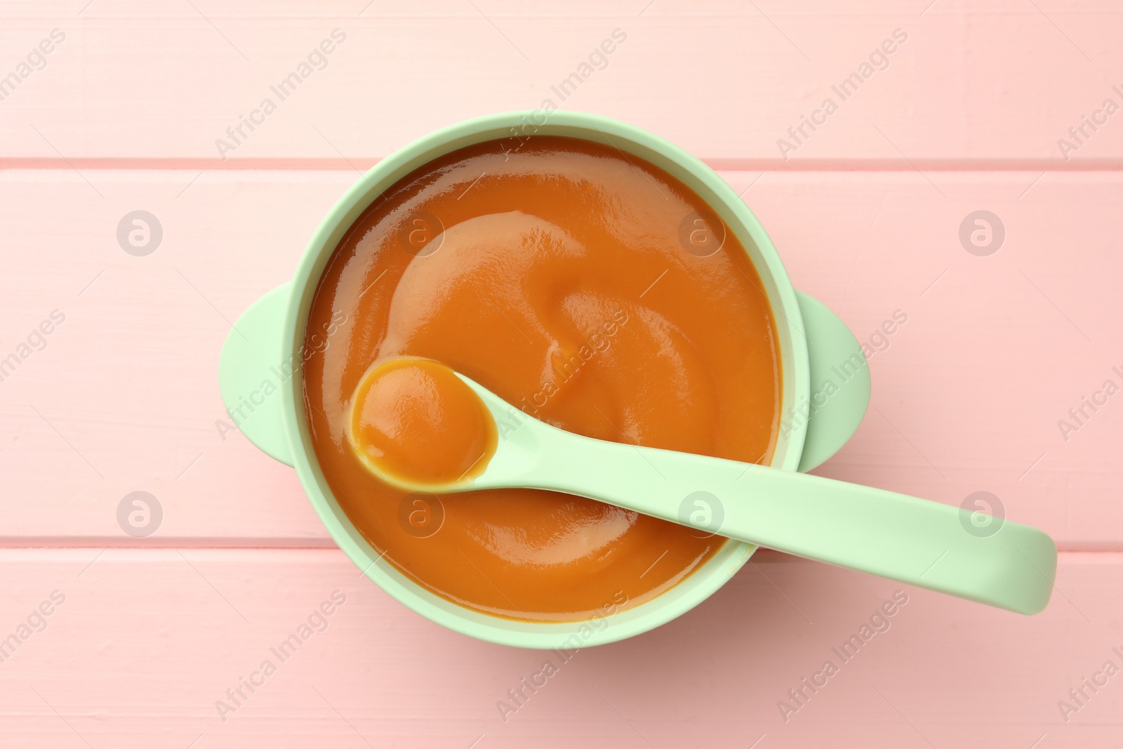 Photo of Bowl and spoon with tasty pureed baby food on pink wooden table, top view