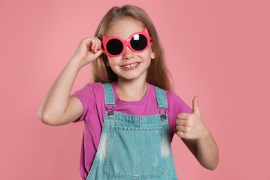 Photo of Smiling girl in stylish sunglasses showing thumb up on pink background