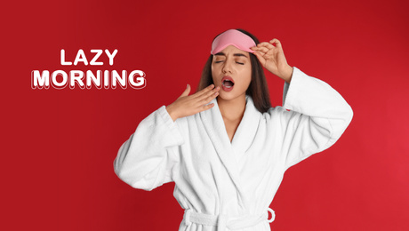 Young woman in bathrobe and eye sleeping mask yawning on red background. Lazy morning