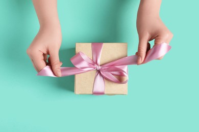 Woman tying bow of gift box on turquoise background, top view