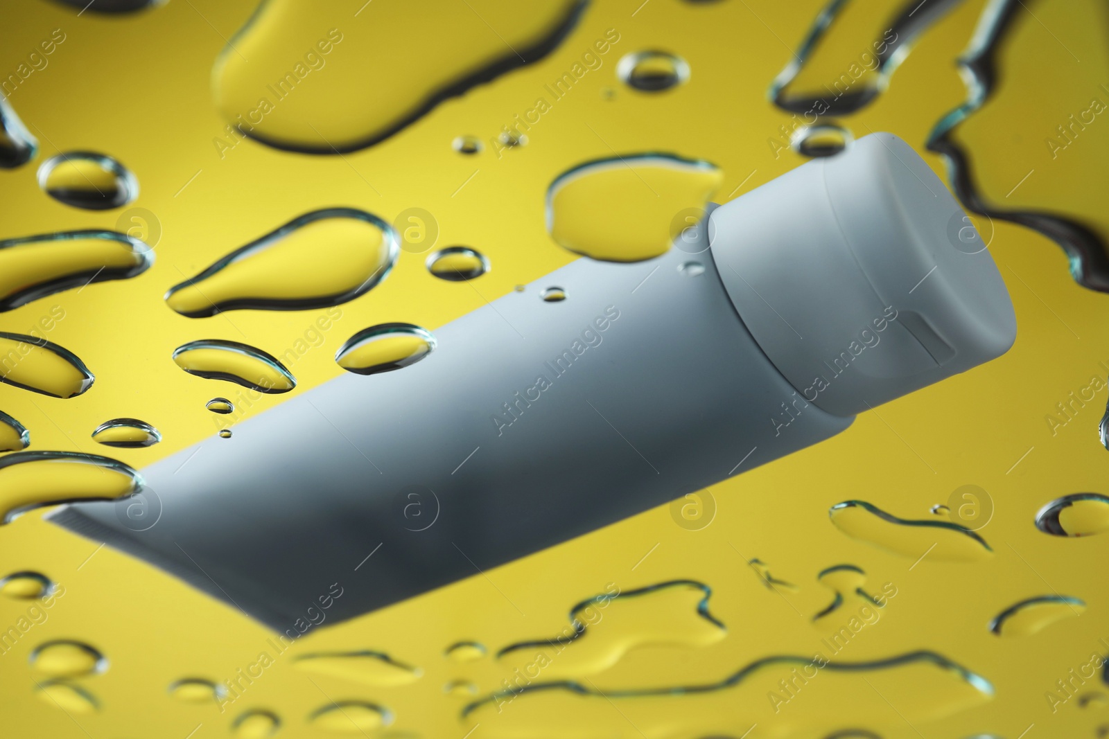 Photo of Moisturizing cream in tube on glass with water drops against yellow background