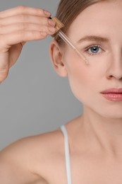 Photo of Beautiful young woman applying essential oil onto face on grey background, closeup