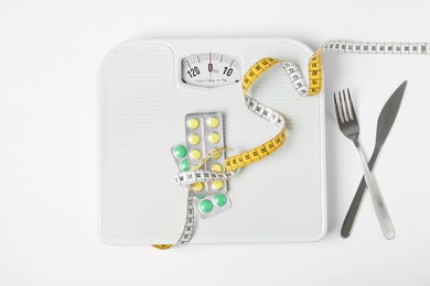 Scales with weight loss pills and measuring tape near cutlery on white background, top view