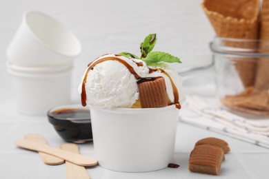 Scoops of tasty ice cream with caramel sauce, mint leaves and candies on white tiled table, closeup