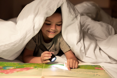 Little boy with flashlight reading book under blanket at home
