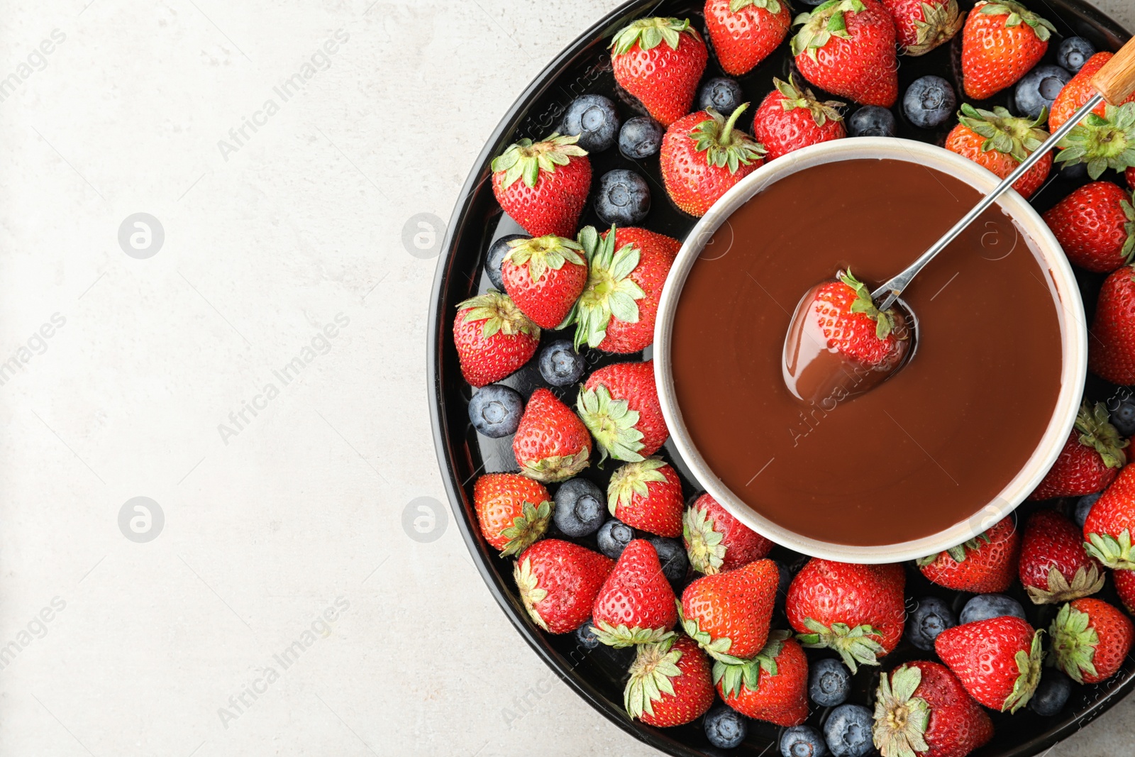 Photo of Fondue fork with strawberry in bowl of melted chocolate surrounded by different berries on light table, top view