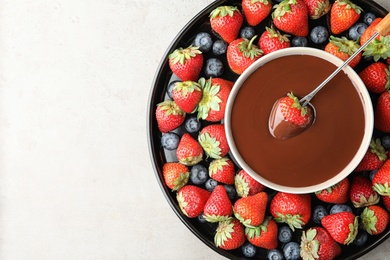 Photo of Fondue fork with strawberry in bowl of melted chocolate surrounded by different berries on light table, top view