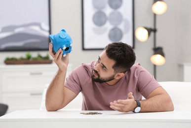 Photo of Worried young man with piggy bank and money at white table indoors