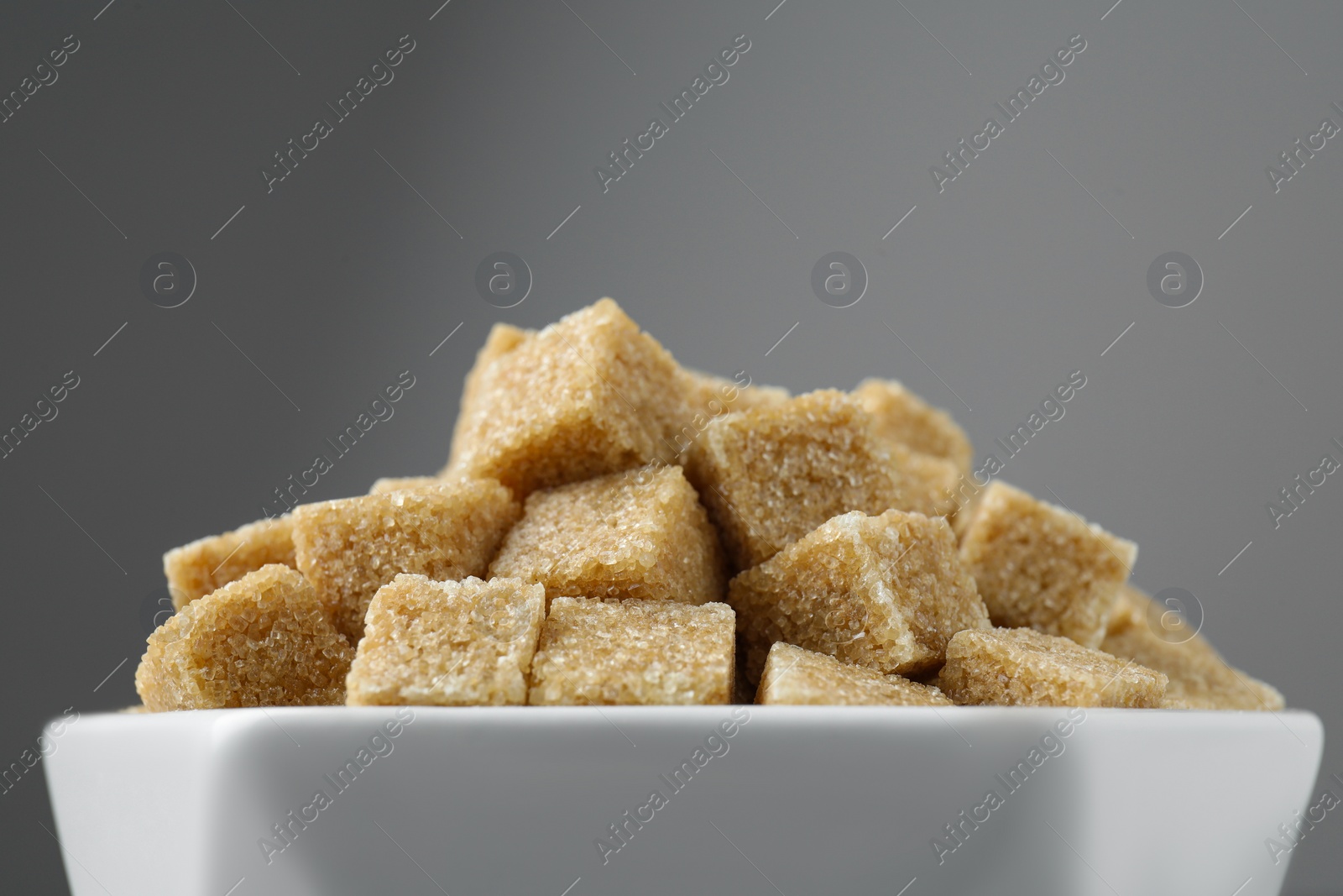 Photo of Brown sugar cubes in bowl on grey background, closeup