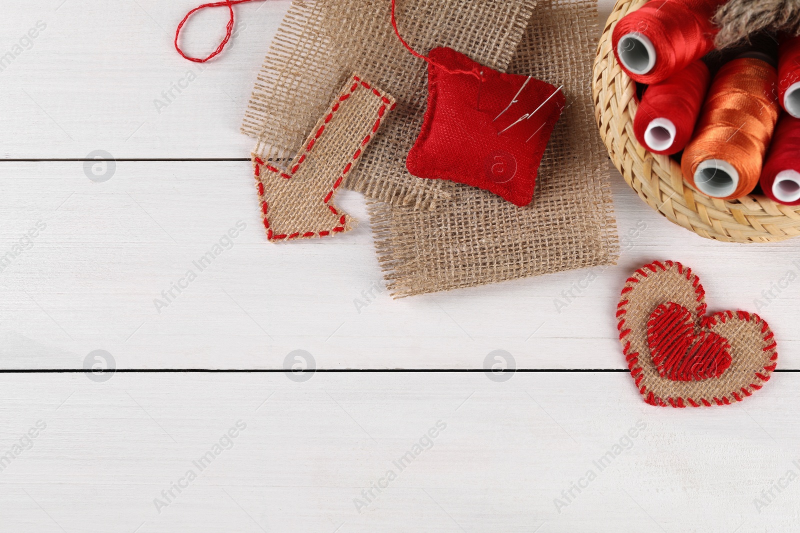 Photo of Pieces of burlap fabric, spools of threads, pincushion and needles on white wooden table, flat lay. Space for text