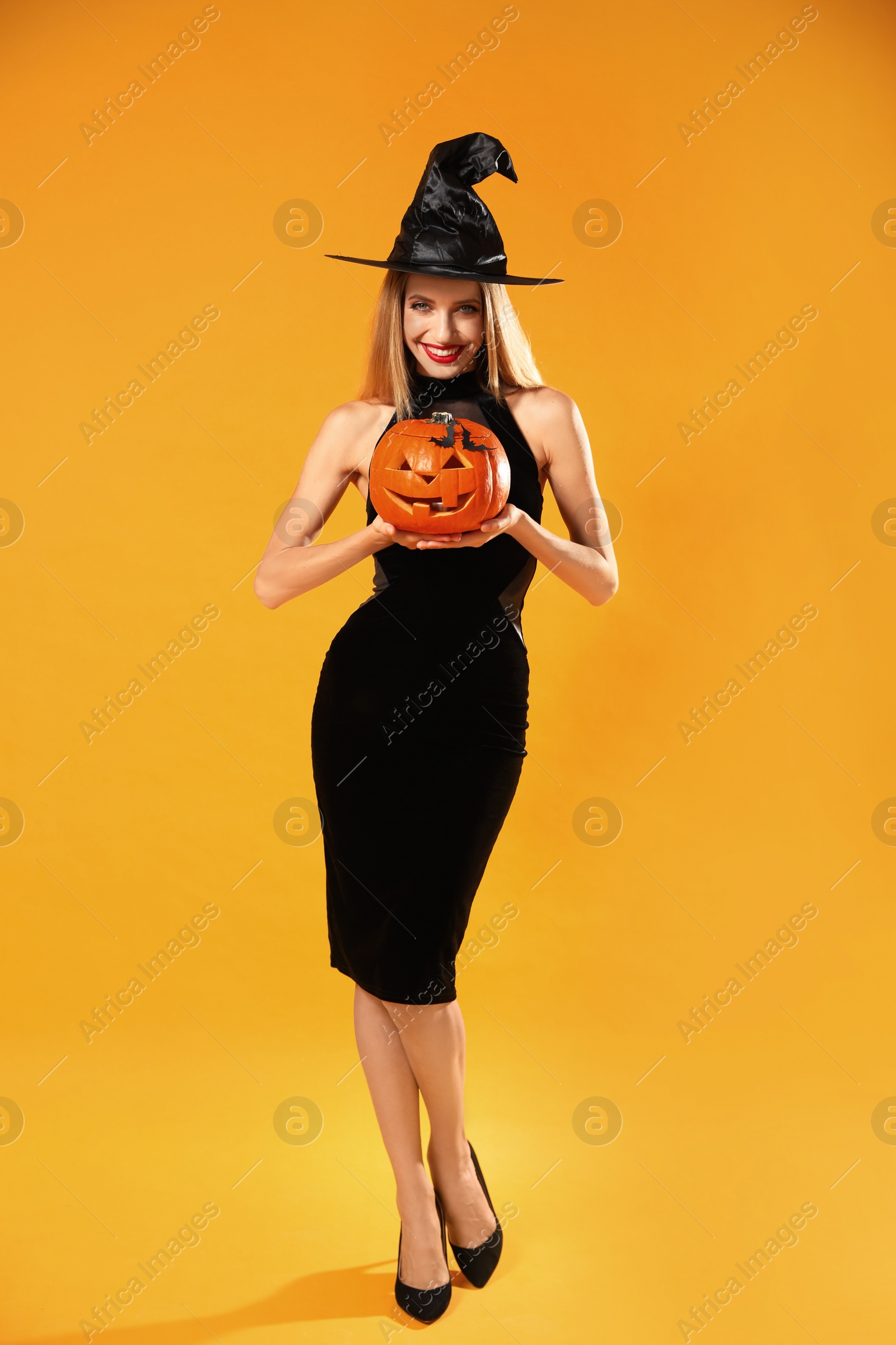 Photo of Beautiful woman in witch costume with jack o'lantern on yellow background. Halloween party