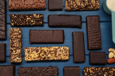 Different tasty bars, nuts and scoop of protein powder on blue wooden table, flat lay