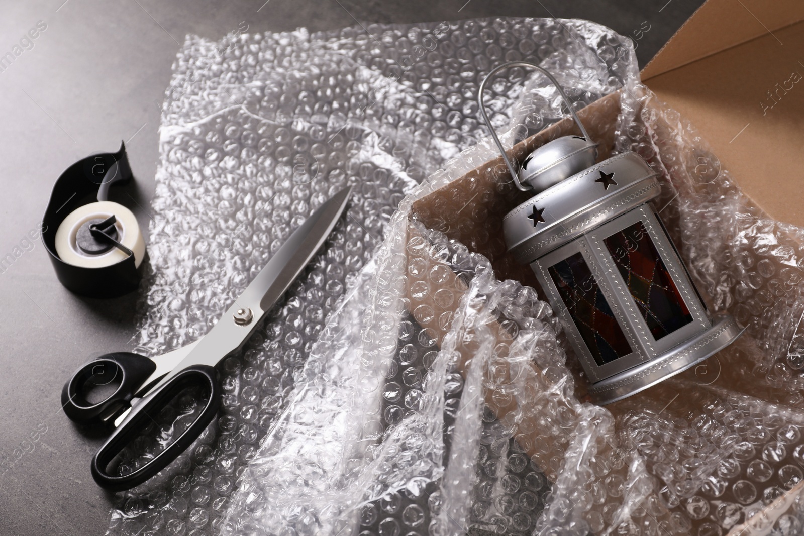 Photo of Beautiful decorative lantern, bubble wrap, scissors and cardboard box on dark grey table