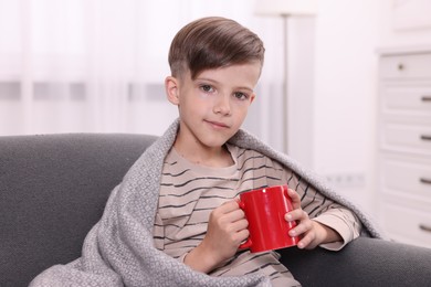 Photo of Cute boy under plaid with red ceramic mug on sofa at home