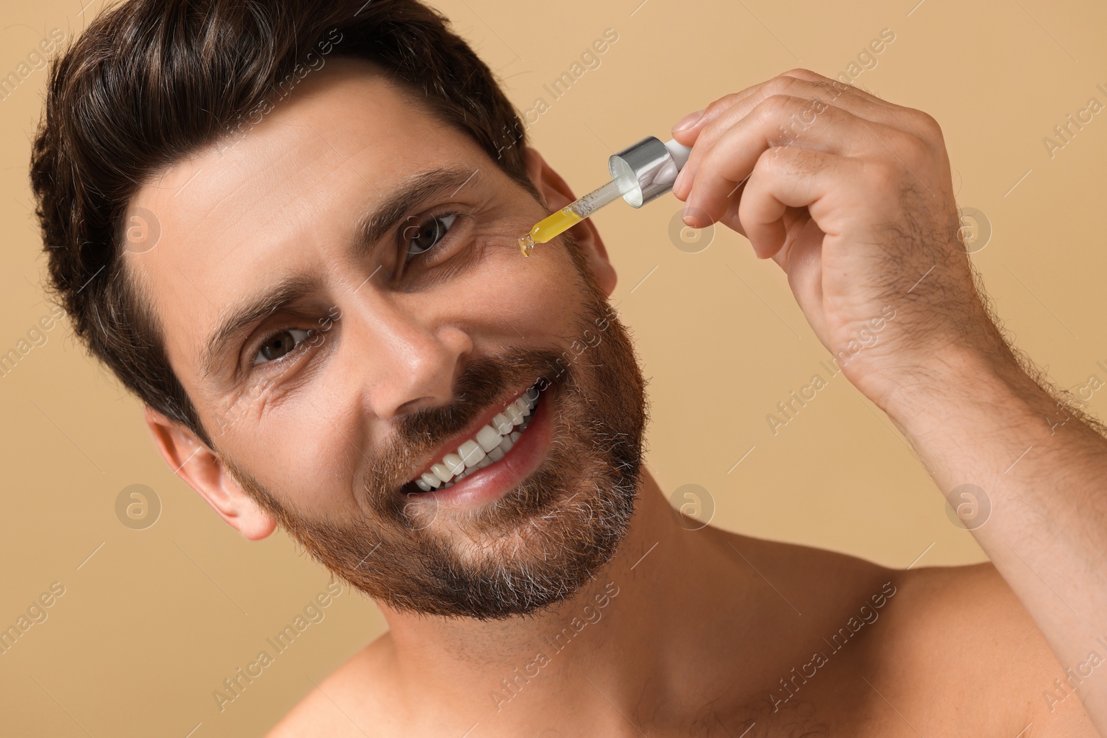 Photo of Smiling man applying cosmetic serum onto his face on beige background, closeup