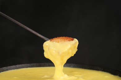 Photo of Dipping piece of bread into fondue pot with tasty melted cheese against dark gray background, closeup