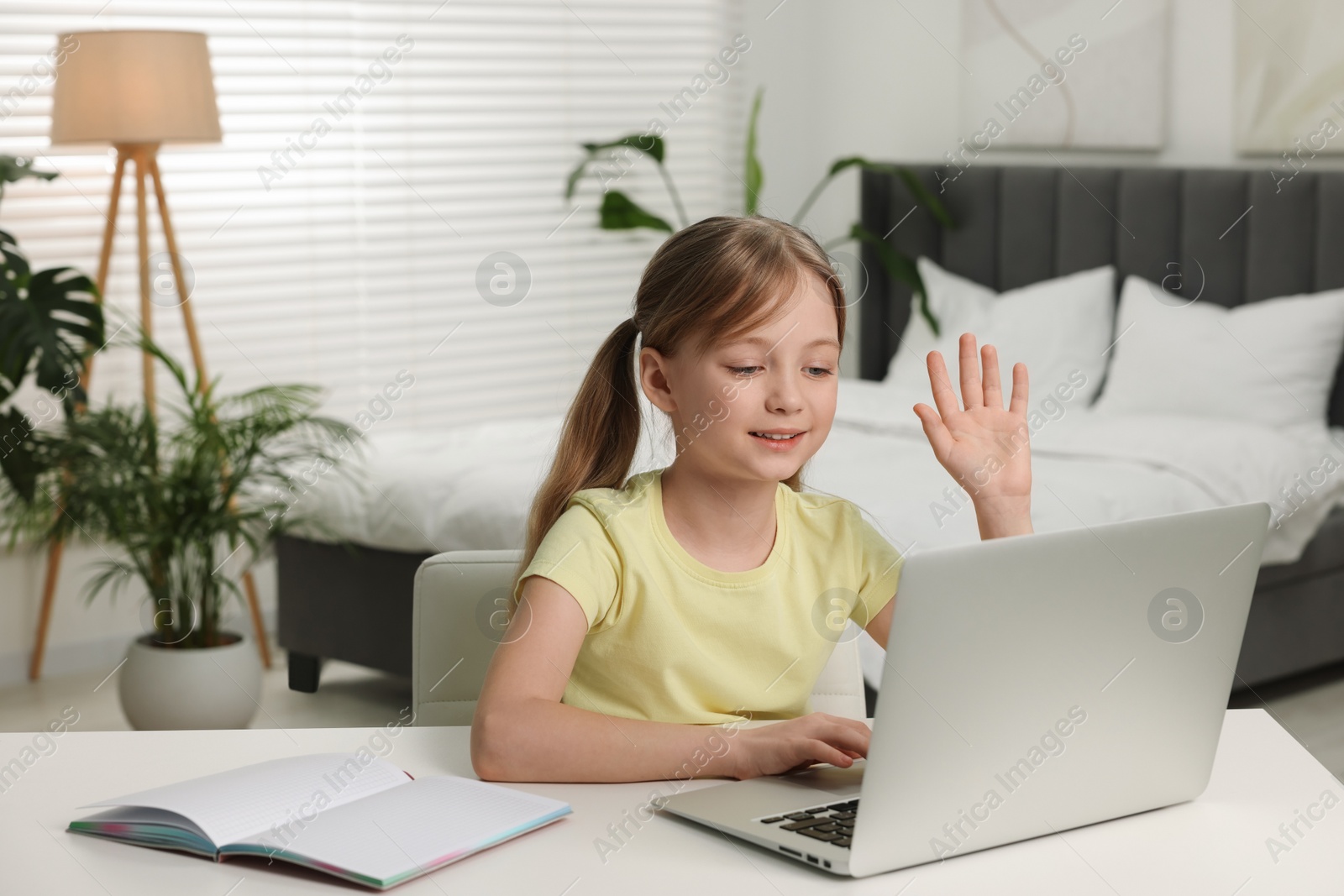 Photo of Cute girl waving hello during online lesson via laptop at white table indoors