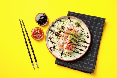 Delicious spring rolls, sauces and chopsticks on yellow background, flat lay