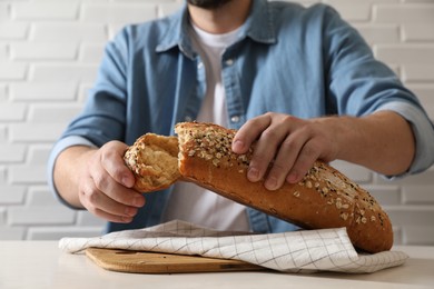 Photo of Man breaking loaf of fresh bread at white table near brick wall, closeup