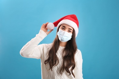 Photo of Beautiful woman wearing Santa Claus hat and medical mask on light blue background