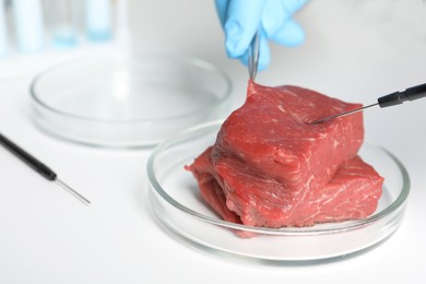 Photo of Scientist with dissecting needle and tweezers examining piece of raw cultured meat at white table, closeup. Space for text