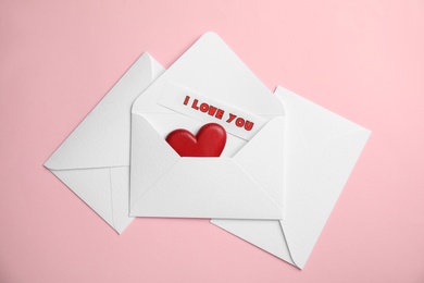 Sheet of paper with phrase I Love You and decorative heart in envelope on pink background, flat lay