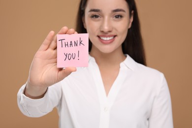 Photo of Happy woman holding paper note with phrase Thank You on brown background, selective focus
