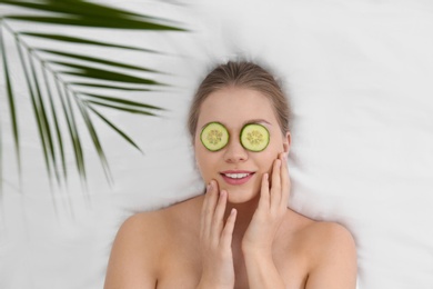 Photo of Beautiful woman with cucumber slices and tropical leaf on white fabric, above view. Organic face mask