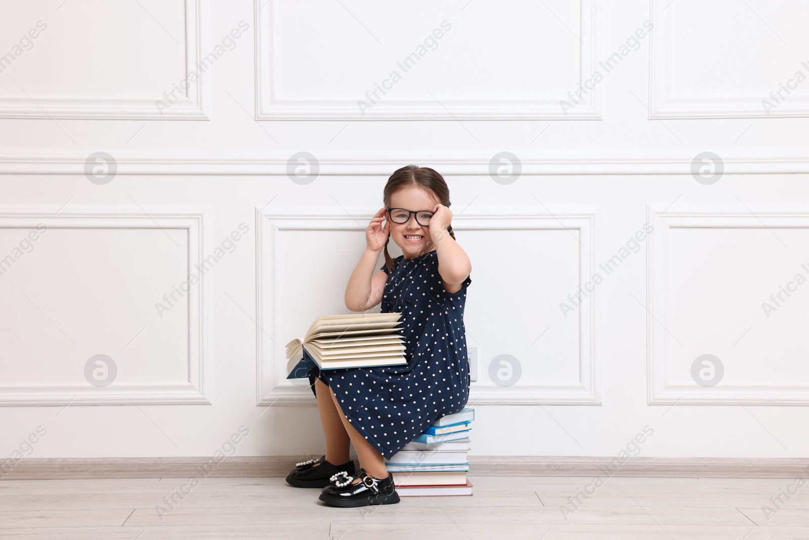 Photo of Cute little girl in glasses sitting on stack of books near white wall