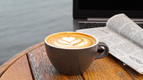 Photo of Cup of delicious coffee, laptop and newspaper on wooden table