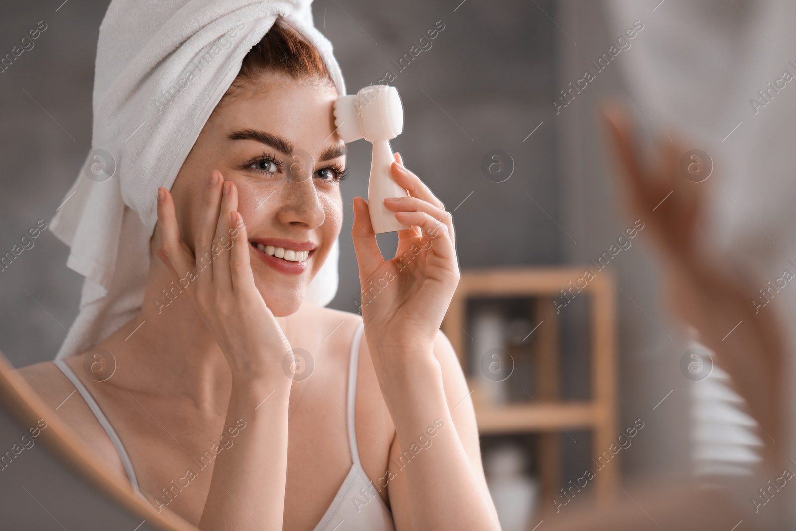 Photo of Washing face. Young woman with cleansing brush near mirror in bathroom