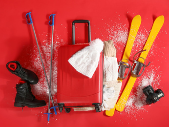 Photo of Suitcase with warm clothes, camera and skis on red background, flat lay. Winter vacation