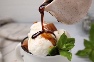 Photo of Pouring caramel sauce onto ice cream with candies and mint leaves, closeup