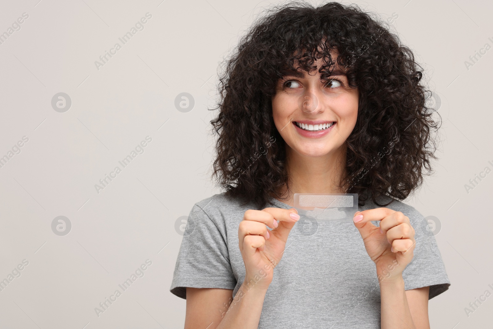 Photo of Young woman holding teeth whitening strips on light grey background, space for text