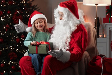 Photo of Merry Christmas. Little girl sitting on Santa's knee and holding gift at home