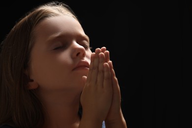 Photo of Cute little girl with hands clasped together praying on black background. Space for text