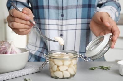 Photo of Woman taking clove of pickled garlic from jar at grey table in kitchen, closeup