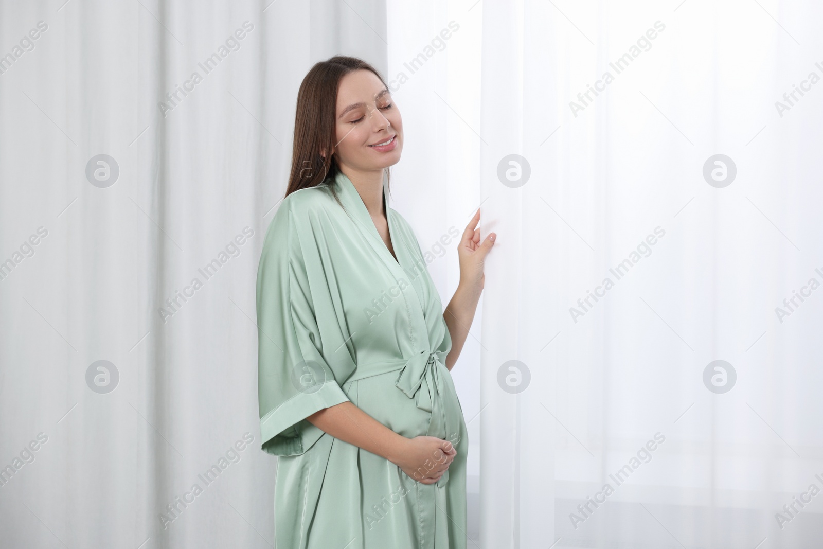 Photo of Beautiful pregnant woman in green dressing gown near window indoors