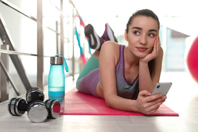 Lazy young woman with smartphone on yoga mat indoors