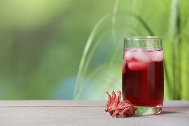 Photo of Refreshing hibiscus tea with ice cubes in glass and roselle flowers on white wooden table against blurred green background. Space for text