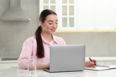 Woman writing something in notebook while using laptop at white table in kitchen