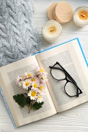 Open book with chamomile flowers as bookmark, scented candles and glasses on white wooden table, flat lay