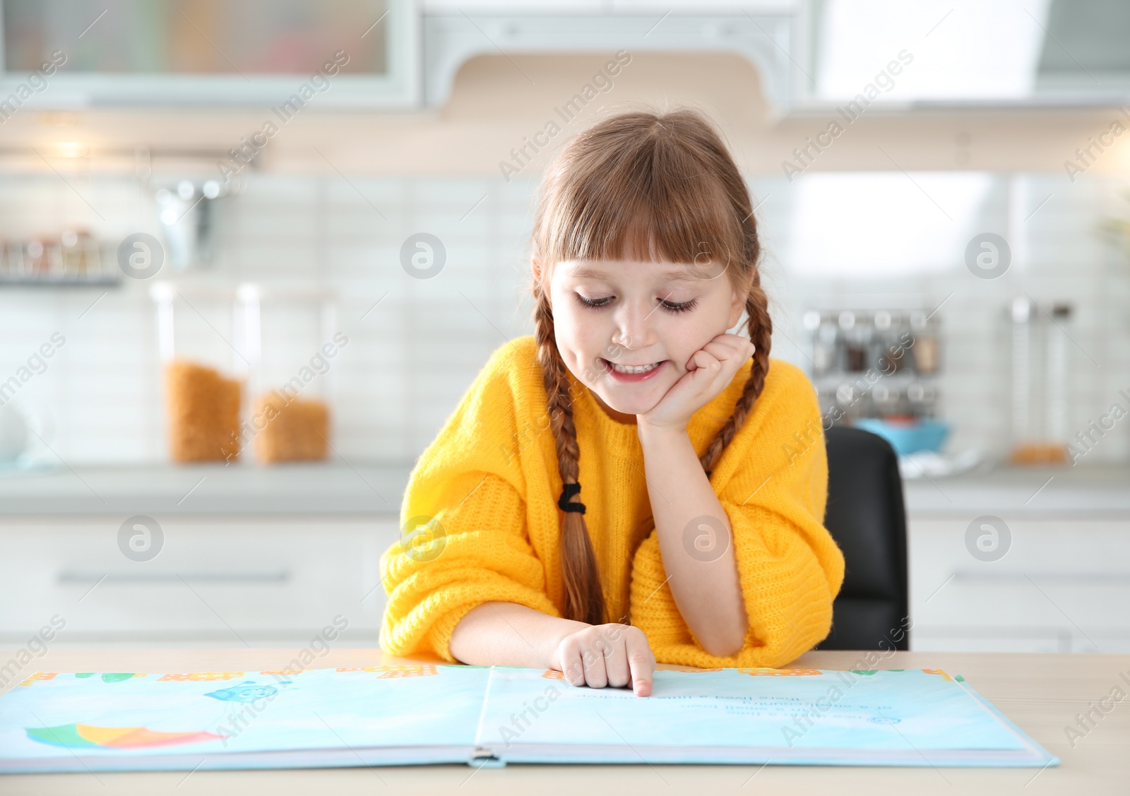 Photo of Cute little girl reading book at table in kitchen