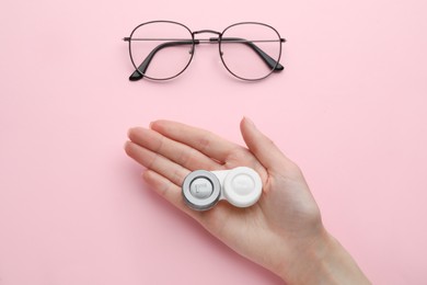 Photo of Woman holding case with contact lenses near glasses on pink background, top view