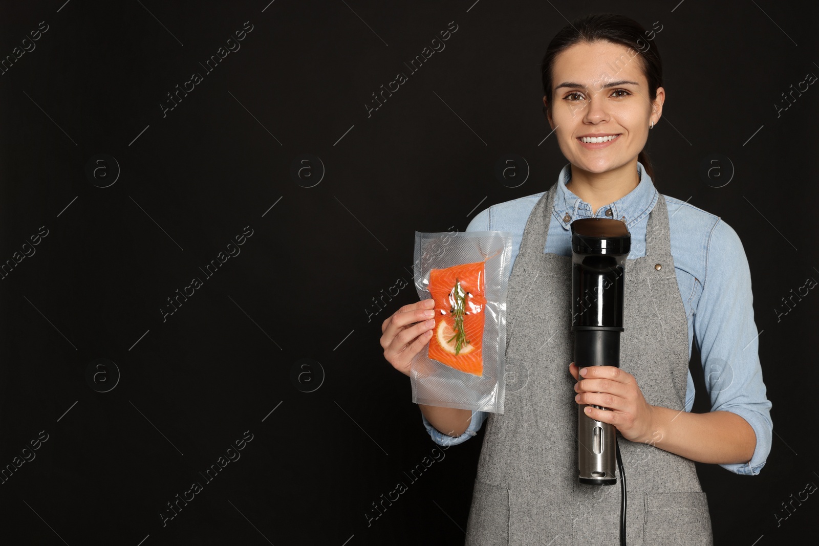 Photo of Beautiful young woman holding sous vide cooker and salmon in vacuum pack on black background. Space for text