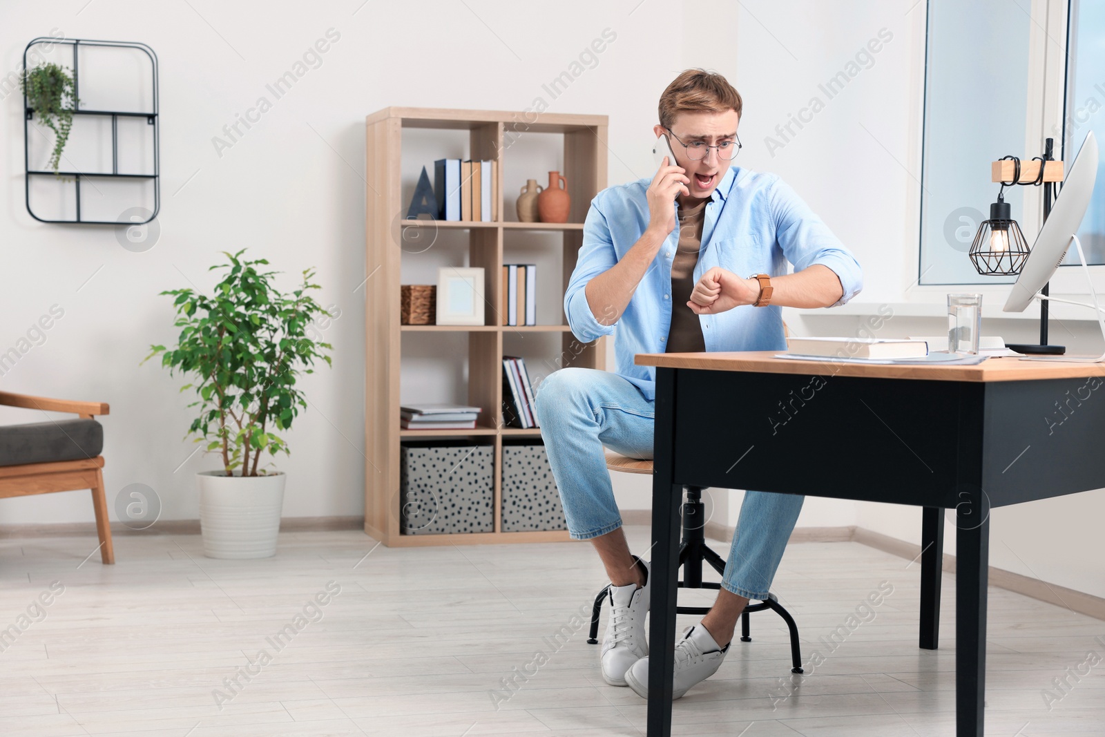 Photo of Emotional young man checking time while talking on phone in office. Being late