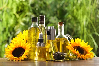 Photo of Many different bottles with cooking oil, sunflower seeds and flowers on wooden table against blurred green background