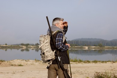 Photo of Man with hunting rifle and backpack near lake outdoors