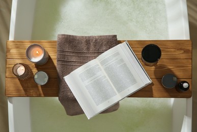 Photo of Wooden bath tray with open book, glass of wine and cosmetic products on tub indoors, top view