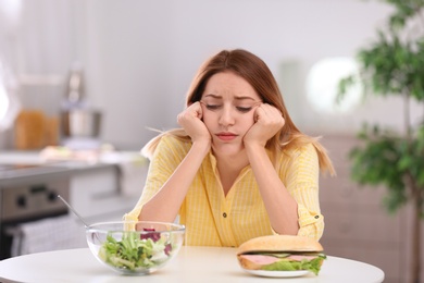 Sad young woman choosing between salad and sandwich in kitchen. Healthy diet